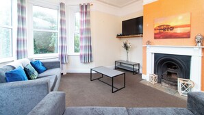 A living room with a grey sofa, a coffee table, a flat-screen TV mounted above a fireplace, and a painting on the wall. - Thornbury Guest House Room 6 (Southampton)