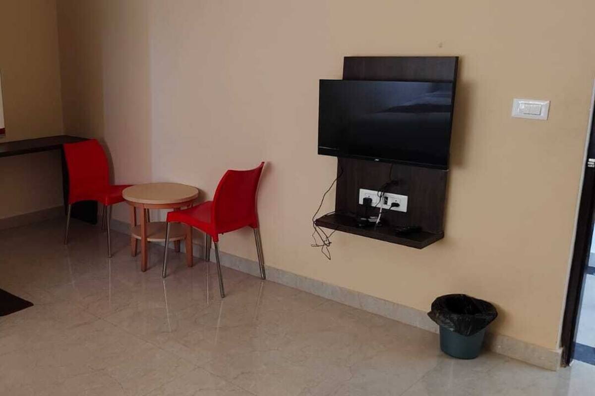 Una habitación de hotel con una cama, un televisor montado en la pared, una mesita redonda con dos sillas rojas y un cubo de basura.