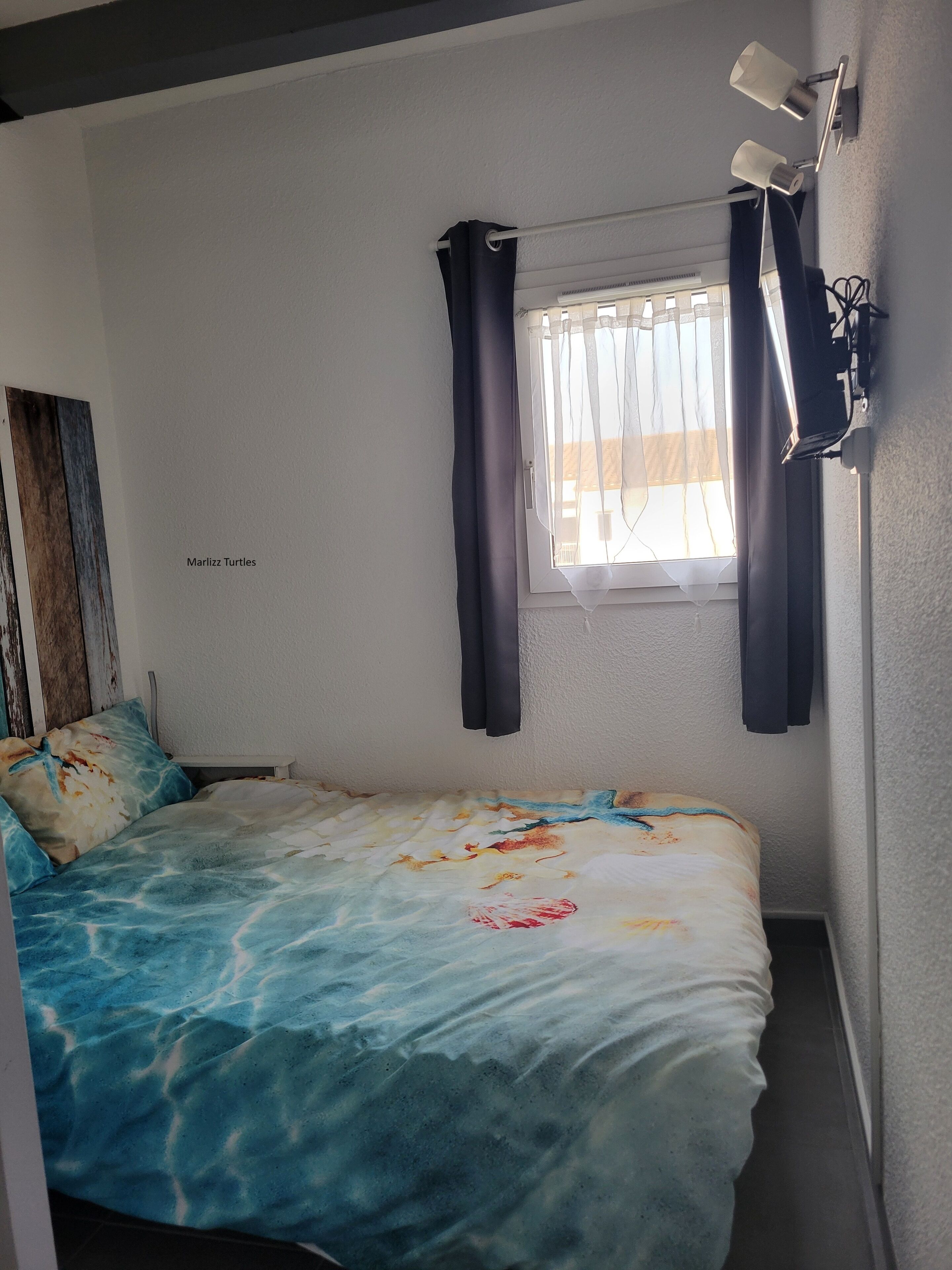 Un dormitorio con cama, ventana con cortinas, lámpara de pared y un cuadro que dice "Marlizz Turtles".