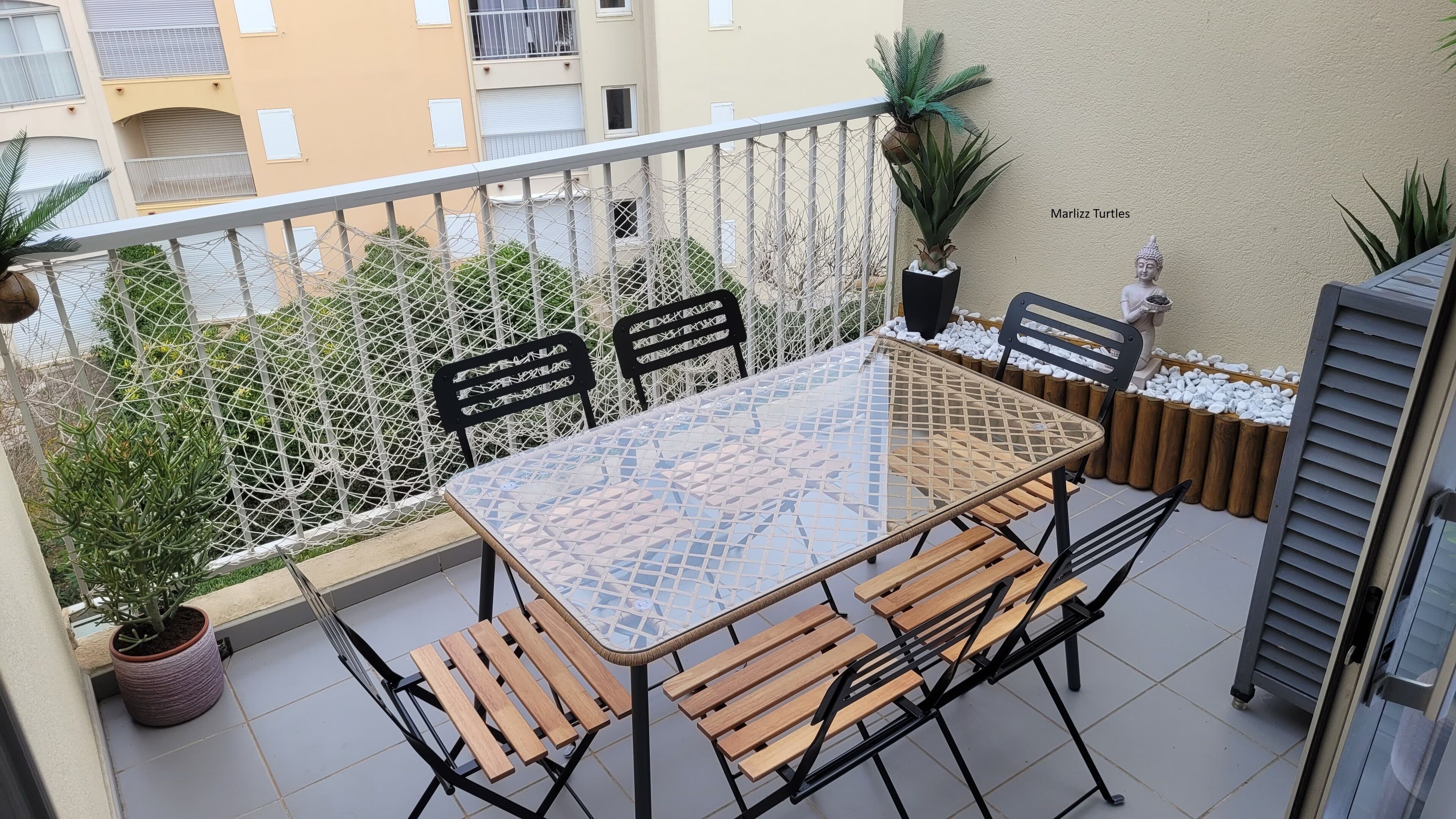 A balcony with a glass-top table, wooden chairs, a white mesh fence, and potted plants.
