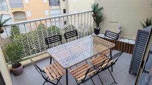 Un balcón con una mesa de vidrio, sillas de madera, una valla de malla blanca y plantas en macetas.