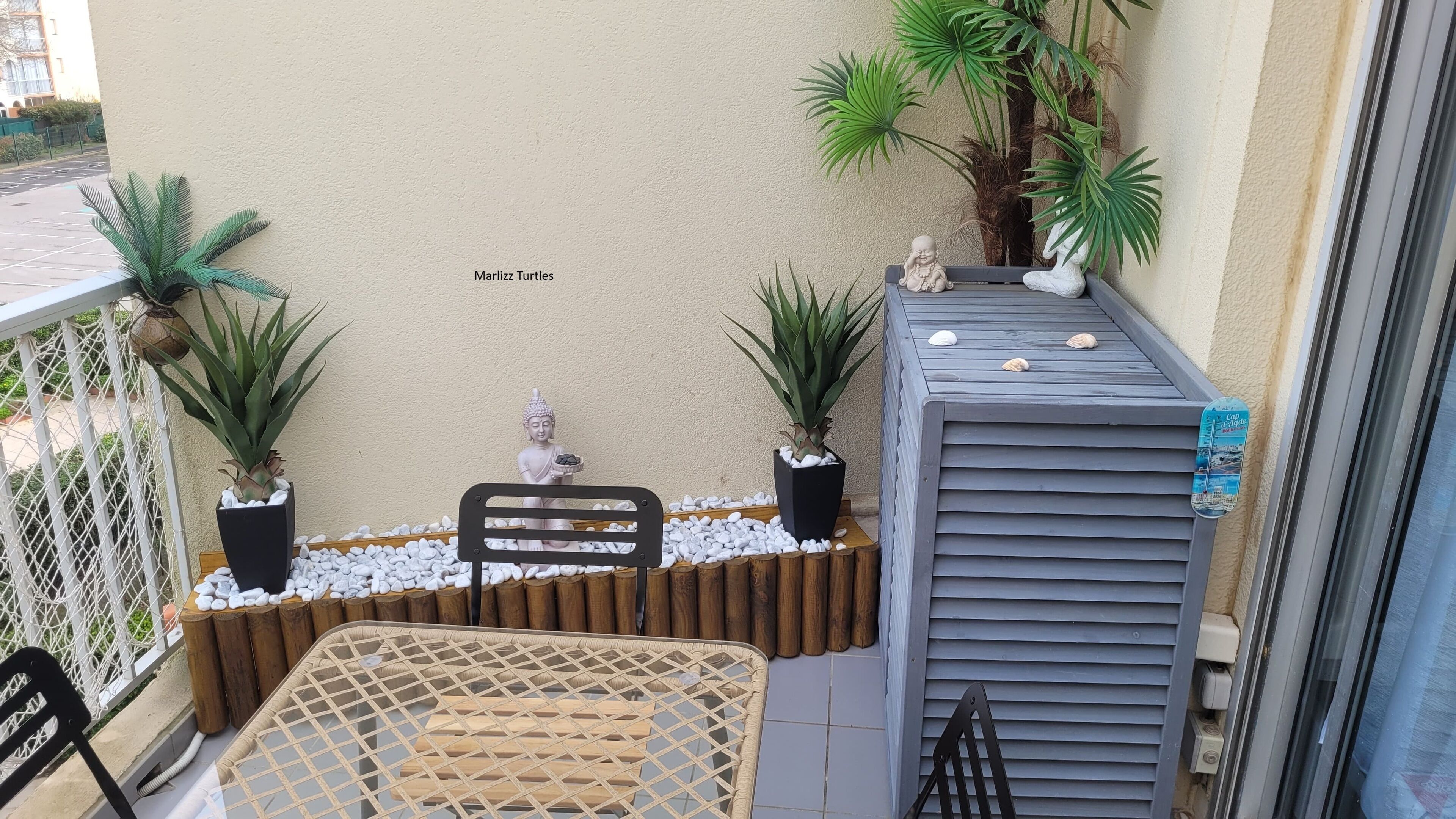 Ein Balkon mit Sitzgelegenheit, zwei Topfpflanzen, einer Buddha-Statue und einem Schild mit der Aufschrift 'Marlizz Turtles'.