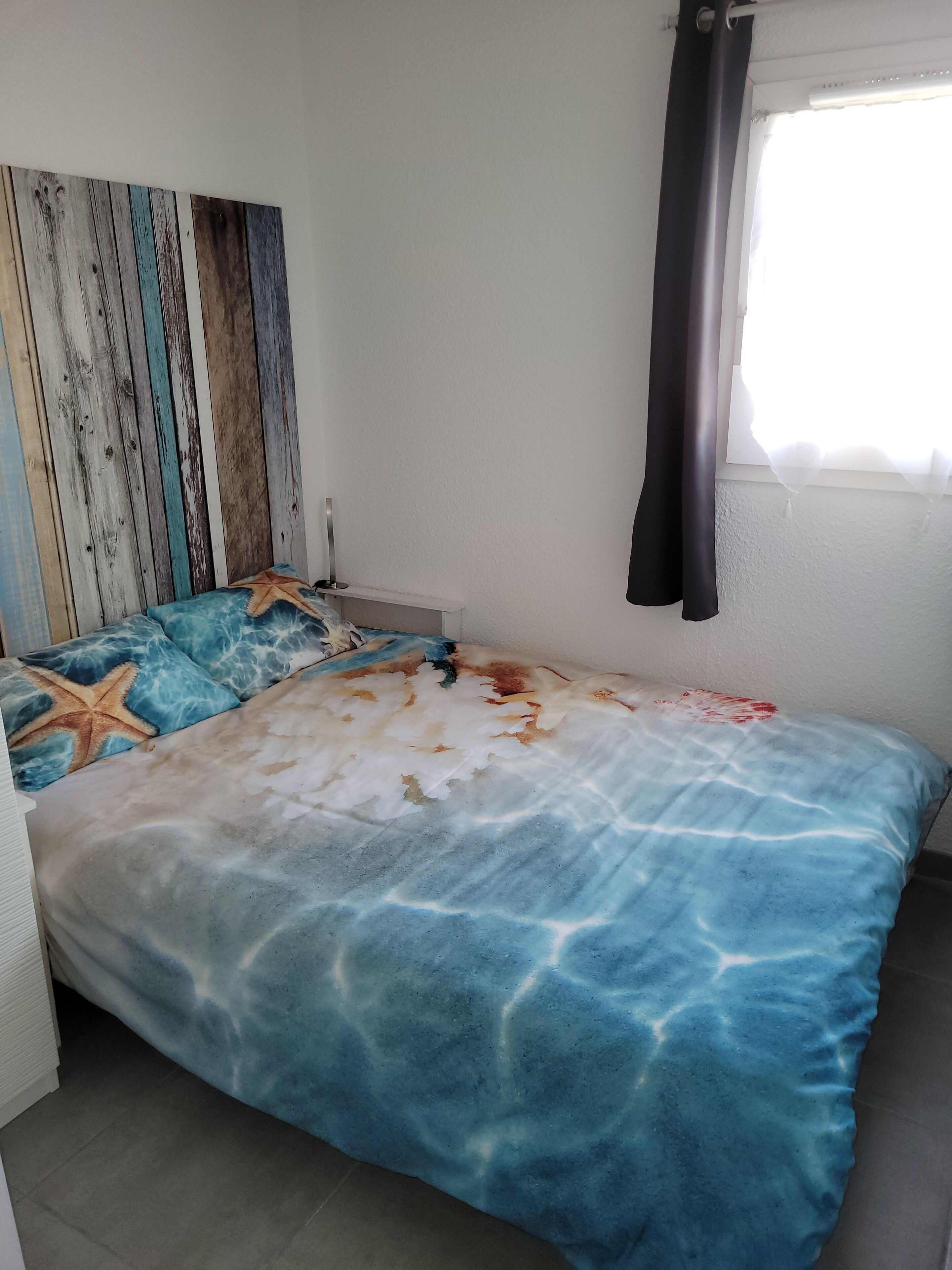 Une chambre à coucher avec un lit agrémenté d’une couette aux motifs bleus et blancs évoquant l’océan, d’un coussin en forme d’étoile de mer et d’une tête de lit en bois.