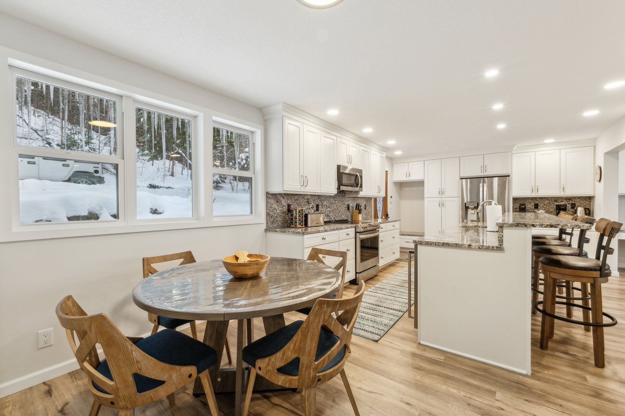 Una cocina moderna con armarios blancos, encimera de granito y comedor con sillas de madera y mesa de vidrio.