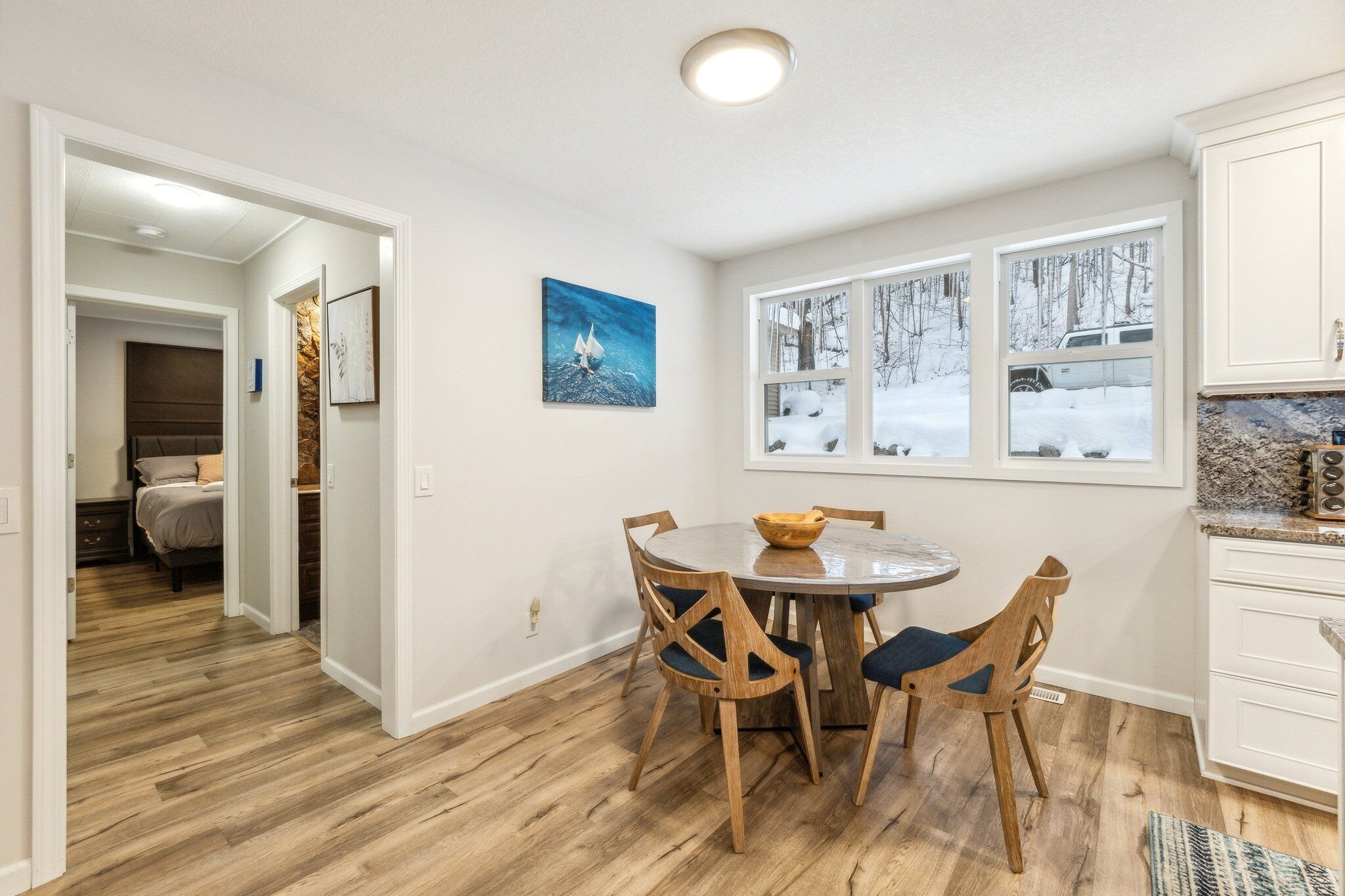 A dining area with a round table and chairs, a window with a snowy view, and a kitchen with white cabinets.