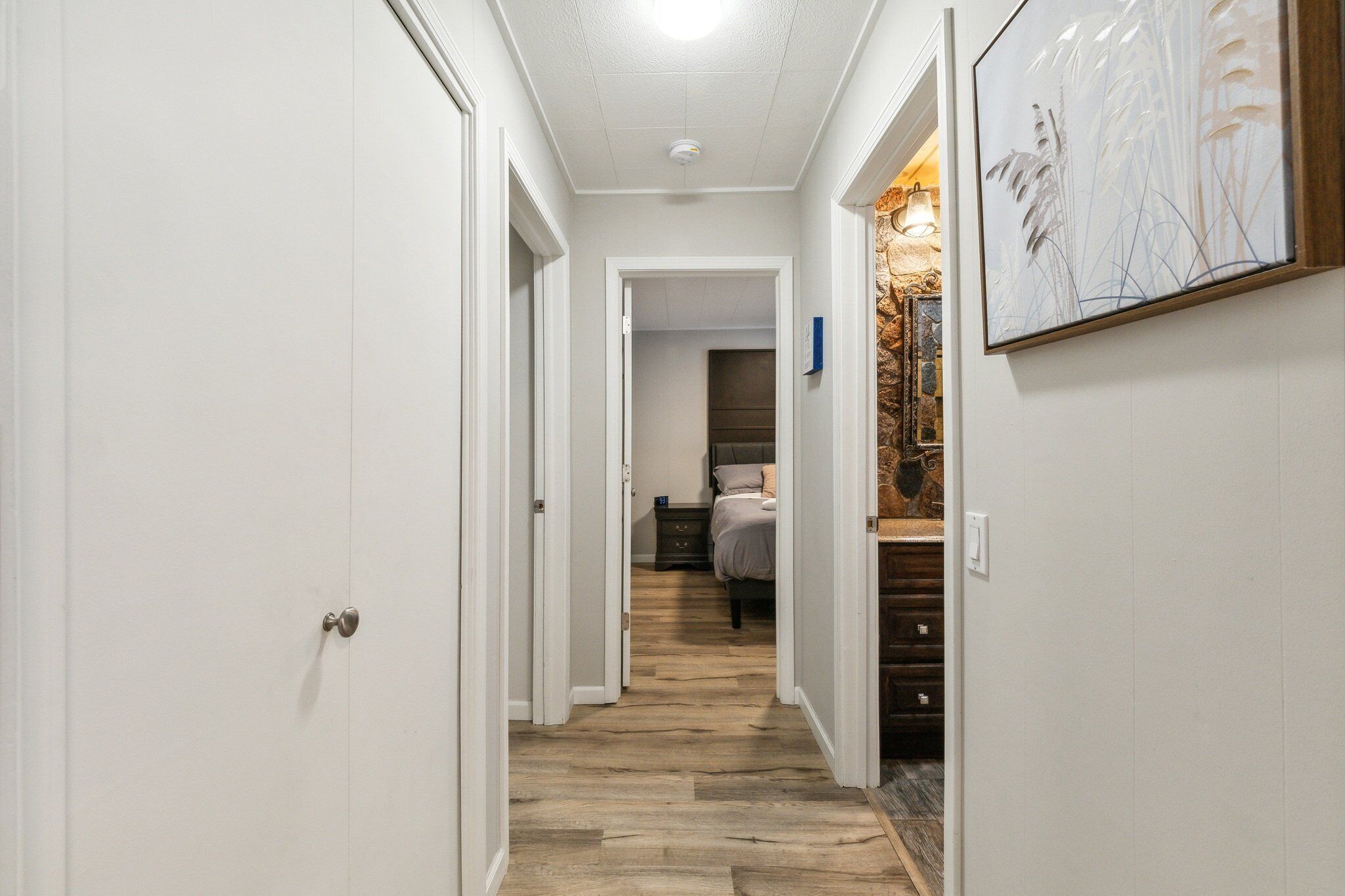A hallway with a wooden floor, white walls, and a bed in the background.