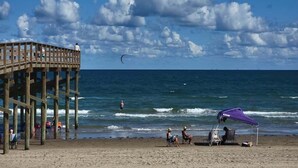 A wooden pier with people on it, a beach with a purple canopy, and the ocean in the background. - Cora Lee Beachview 2 Bedroom Condo (South Padre Island)