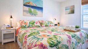 A bedroom with a floral bedspread, a painting of a beach scene, and a window with a view of the water. - Cora Lee Beachview 2 Bedroom Condo (South Padre Island)