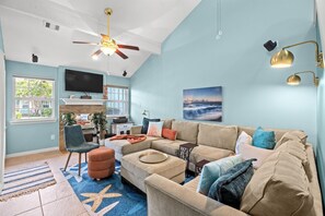 A spacious living room with a large sofa, ottoman, and a television mounted on the wall. - Mermaids Oasis 3 Bedroom Townhouse (Galveston)