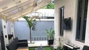 Une terrasse avec un revêtement de sol en gazon synthétique, des meubles en rotin noir et un téléviseur à écran plat fixé au mur.