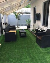 Un patio con césped artificial, mobiliario de mimbre negro y un televisor de pantalla plana montado en la pared.