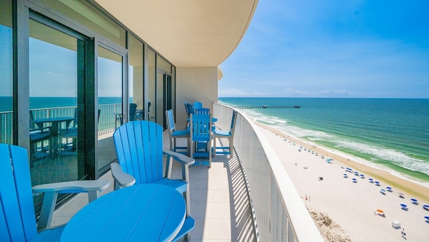 A balcony with blue chairs and tables overlooking a beach with umbrellas and a pier. - Phoenix Gulf Shores Ii 1604 3 Bedroom Condo (Gulf Shores)