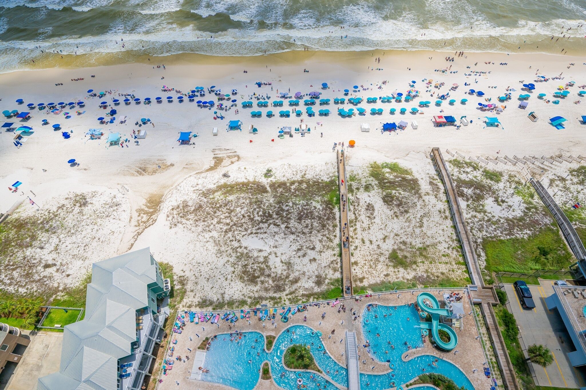 An aerial view of a beach resort with a large pool area, beach umbrellas, and a boardwalk leading to the beach.