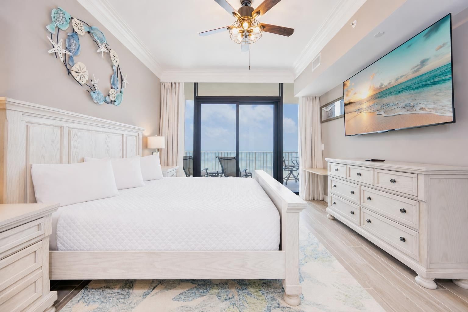A hotel room with a large bed, a ceiling fan, a TV, and a balcony with a view of the ocean.