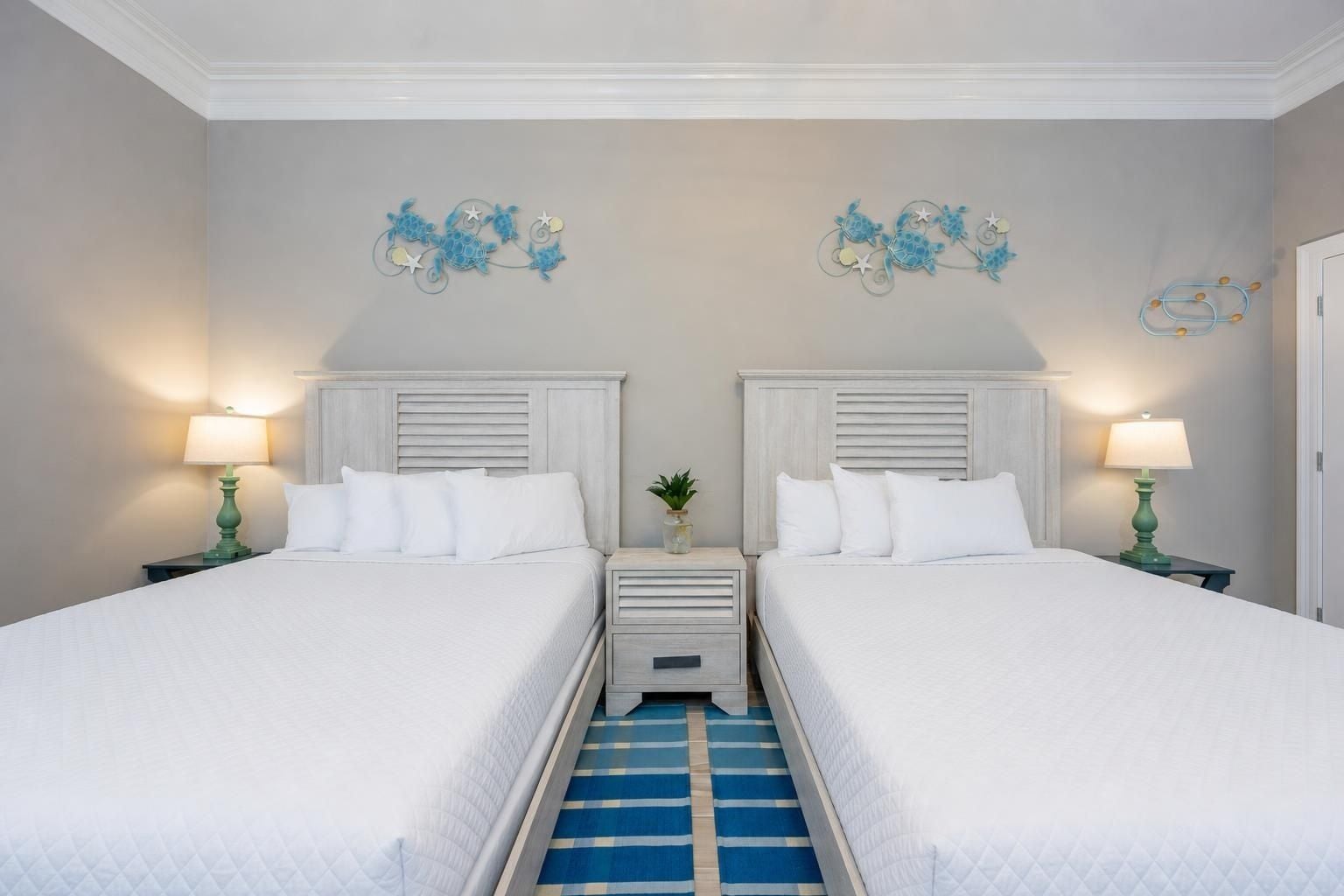 Ein Hotelzimmer mit zwei Betten, jeweils mit weißer Bettwäsche und Kopfteilen, an den Seiten Nachttische mit Lampen. Auf dem Boden ist ein blau-weiß gestreifter Teppich zu sehen.