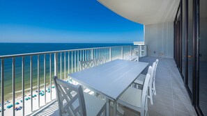 Ein Balkon mit weißem Tisch und Stühlen, der Blick auf einen Strand mit Sonnenschirmen und Menschen f ällt.
