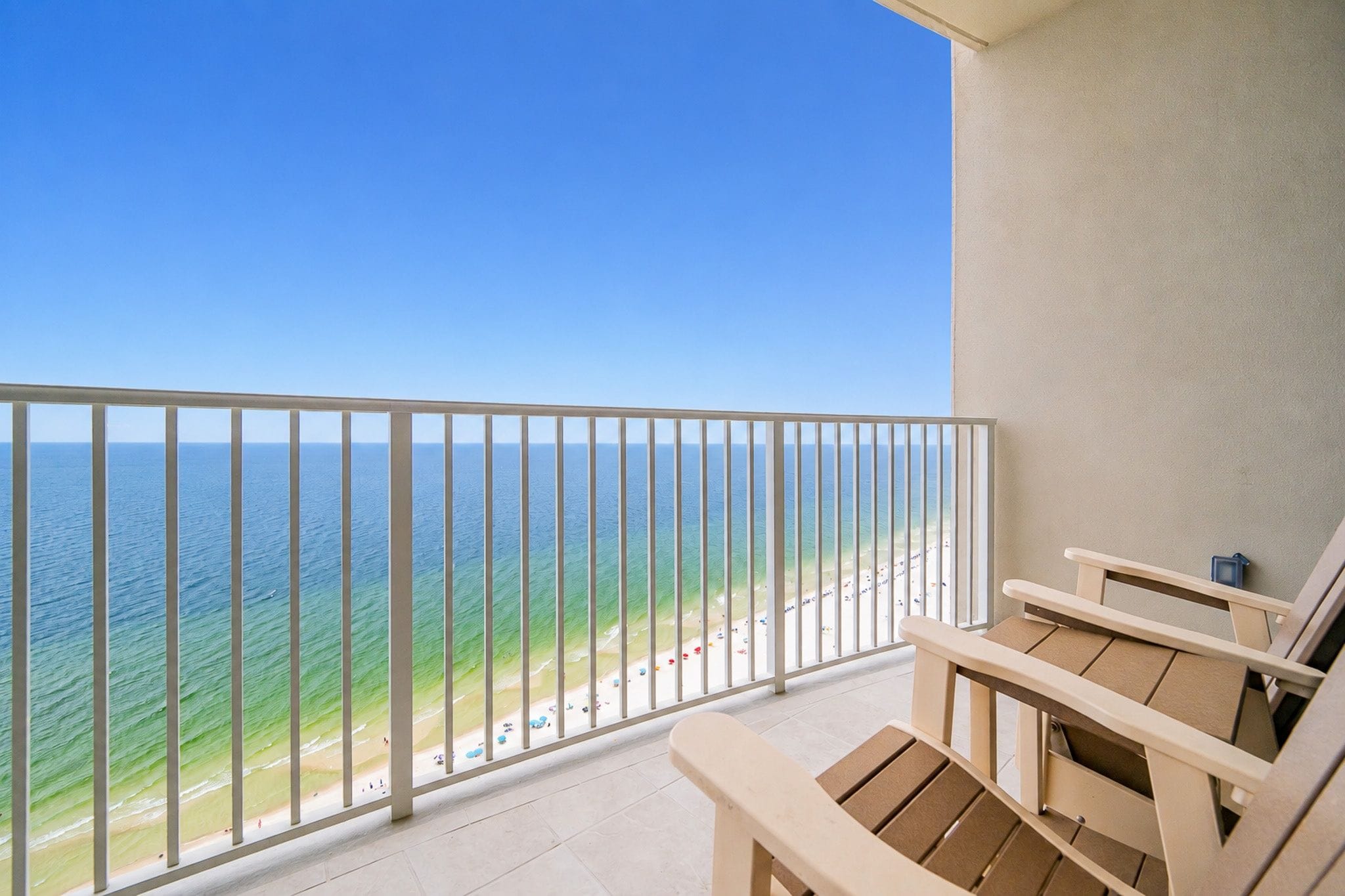A balcony with a view of the ocean and beach, wooden chairs, and a tiled floor.