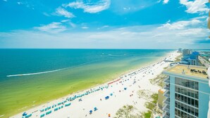 A beach with umbrellas and people, a clear sky, and a building with balconies. - Phoenix Gulf Shores Ii 2303 3 Bedroom Condo (Gulf Shores)