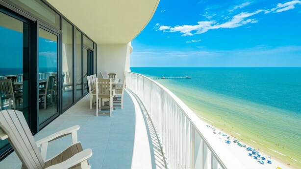 A balcony with white chairs and tables overlooking a beach with clear blue water and a pier in the distance.