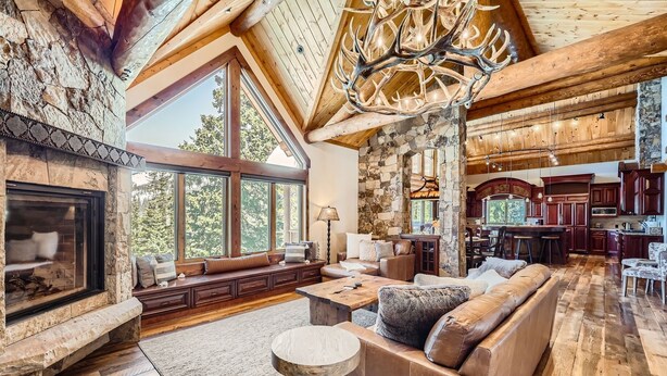 A spacious living room with a stone fireplace, large windows, a high ceiling with wooden beams, and a rustic chandelier. - Bridge To Solitude 6 Bedroom Home (Breckenridge)