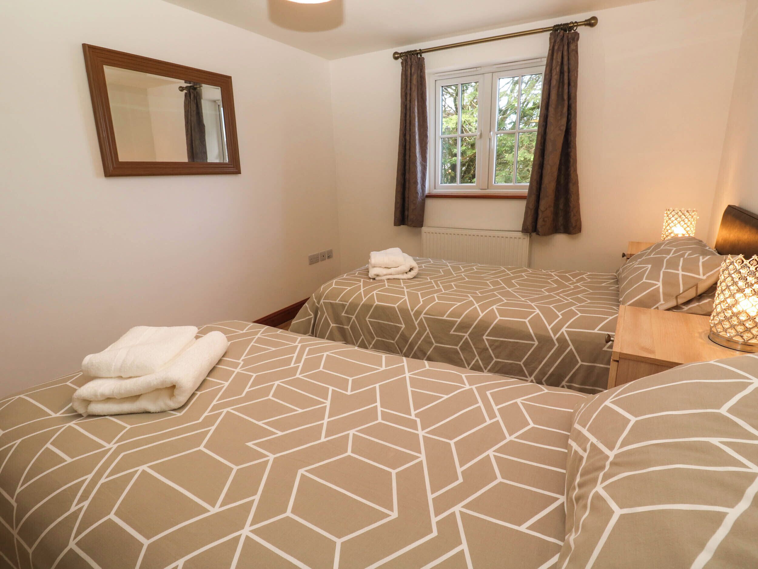 A bedroom with two beds, a window with curtains, a bedside lamp, and a mirror on the wall.