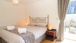 A neatly made bed with white linens and a brown bedspread, a bedside table with a lamp, and a view of a patio area through a window. - The Baler House (Hayle)