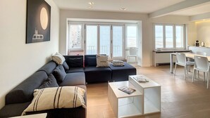 Ein modernes Wohnzimmer mit einem Ecksofa, einem kleinen Couchtisch und einem Essbereich mit weißem Tisch und Stühlen.
