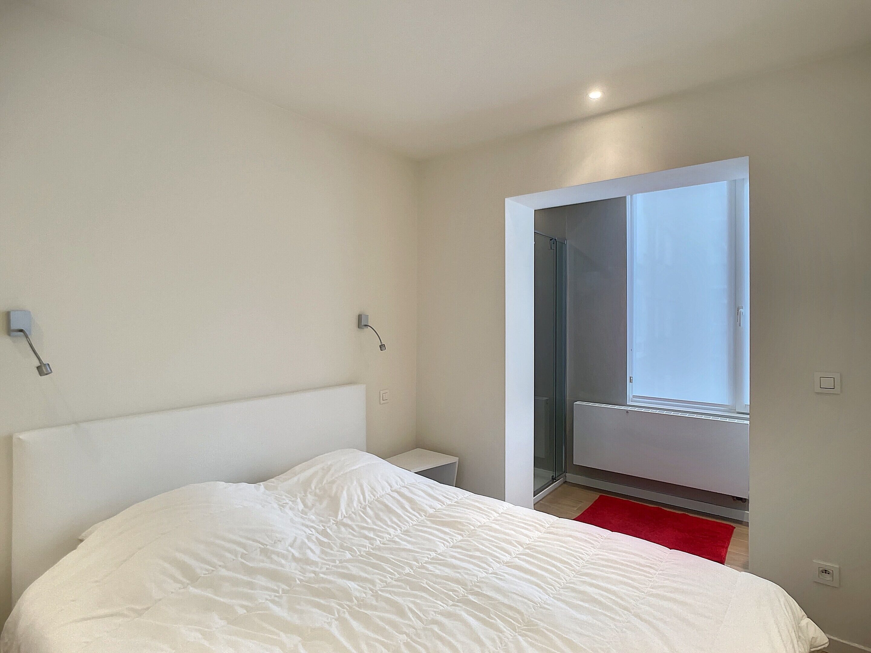 Une chambre avec un grand lit, une table de chevet, un tapis rouge et une porte-fenêtre coulissante donnant sur un balcon.