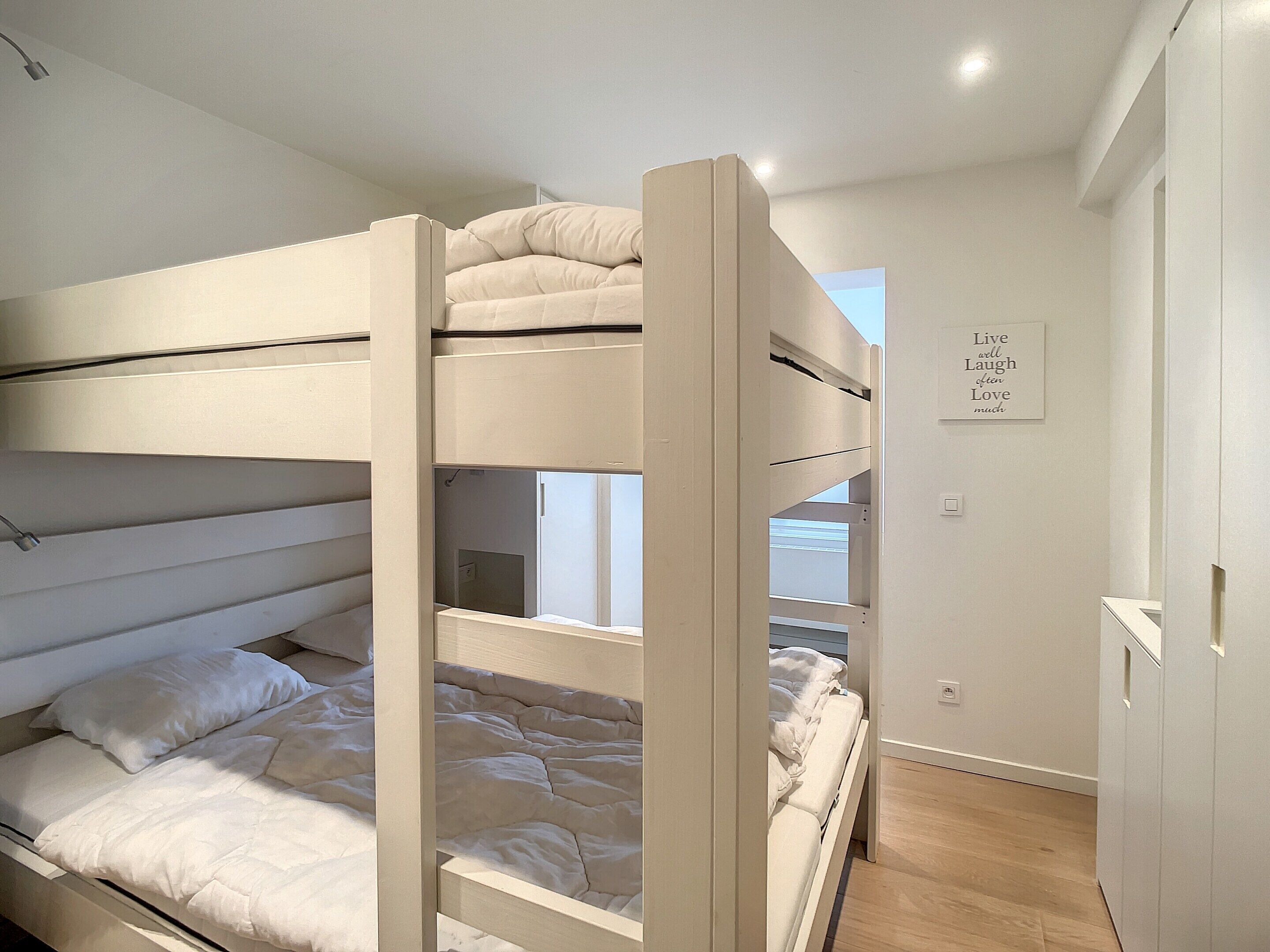 Une chambre avec des lits superposés, décorée dans des tons de blanc, un mur orné d’une citation inspirante et un plancher en bois.