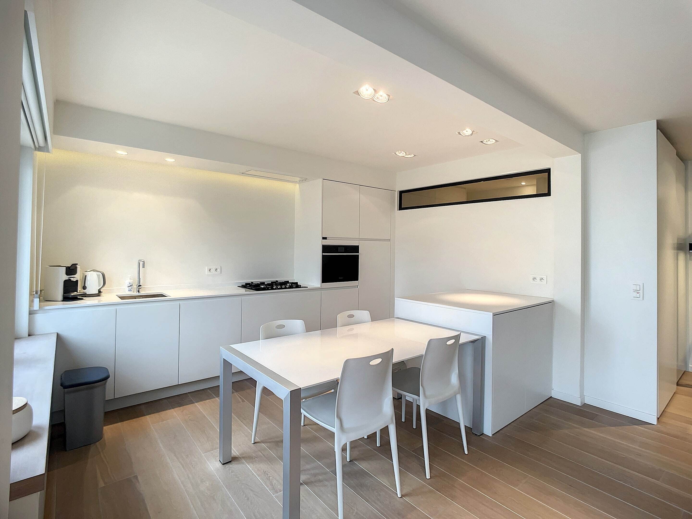 A modern kitchen with white cabinetry, a dining table with chairs, and a small trash bin.