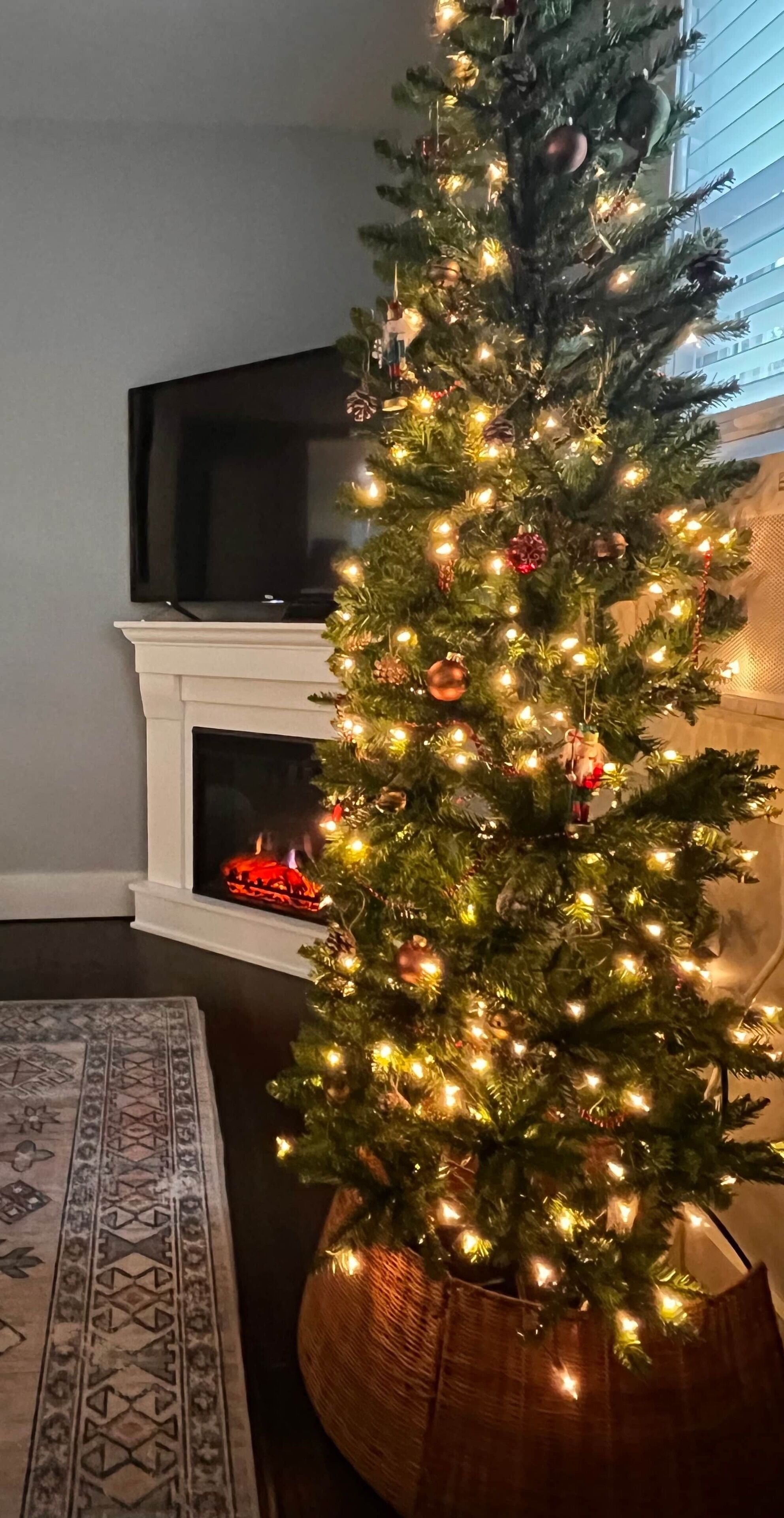 Ein Weihnachtsbaum mit Lichtern und Schmuck, platziert neben einem Kamin und einem Fernseher.