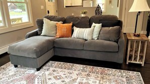 A modern living room with a grey sectional sofa, patterned rug, and a small wooden side table. - Bright, Spotless Cottage Near Bethel Woods (Highland)