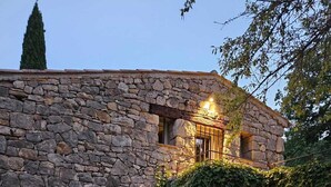 A stone building with a small window and a door, surrounded by greenery and a gravel path.