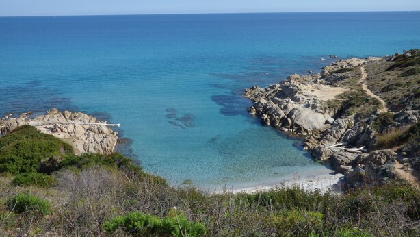 A coastal landscape with clear blue waters, rocky terrain, and a dirt path leading to a sandy beach.