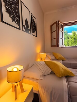 Un dormitorio con una cama, una mesita de noche con lámpara y una ventana con vistas a zonas verdes y montañas.