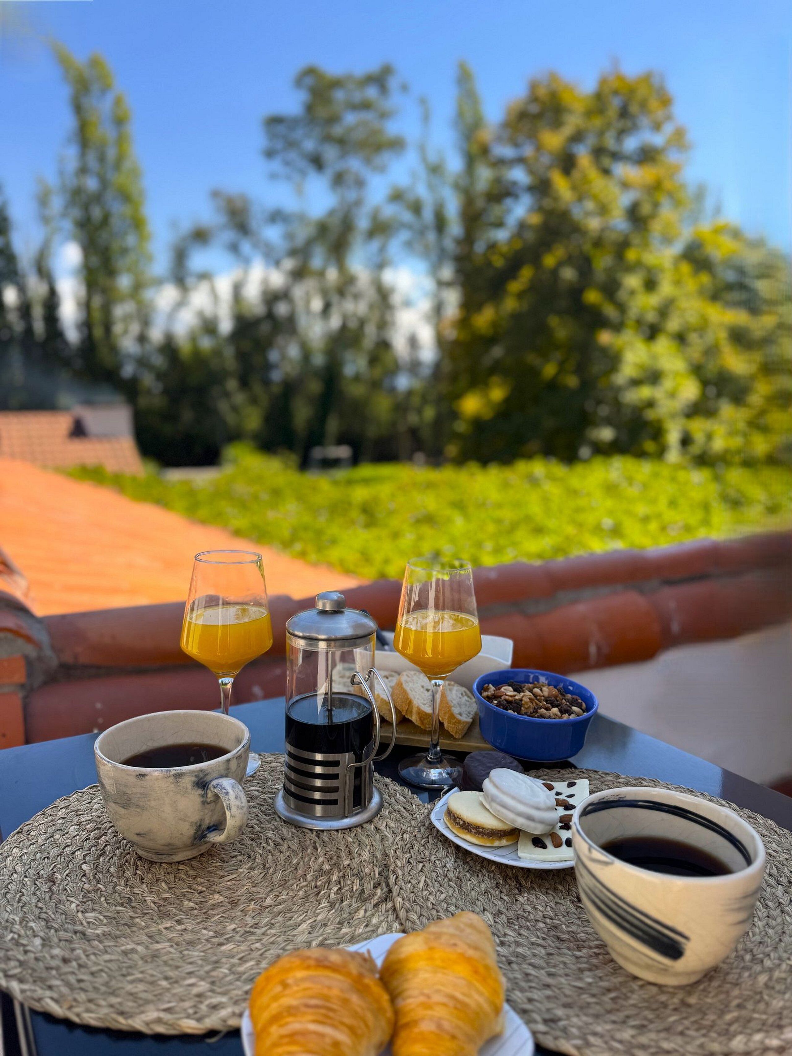 Desayuno dispuesto en una mesa al aire libre con vista a árboles y un muro de ladrillo.