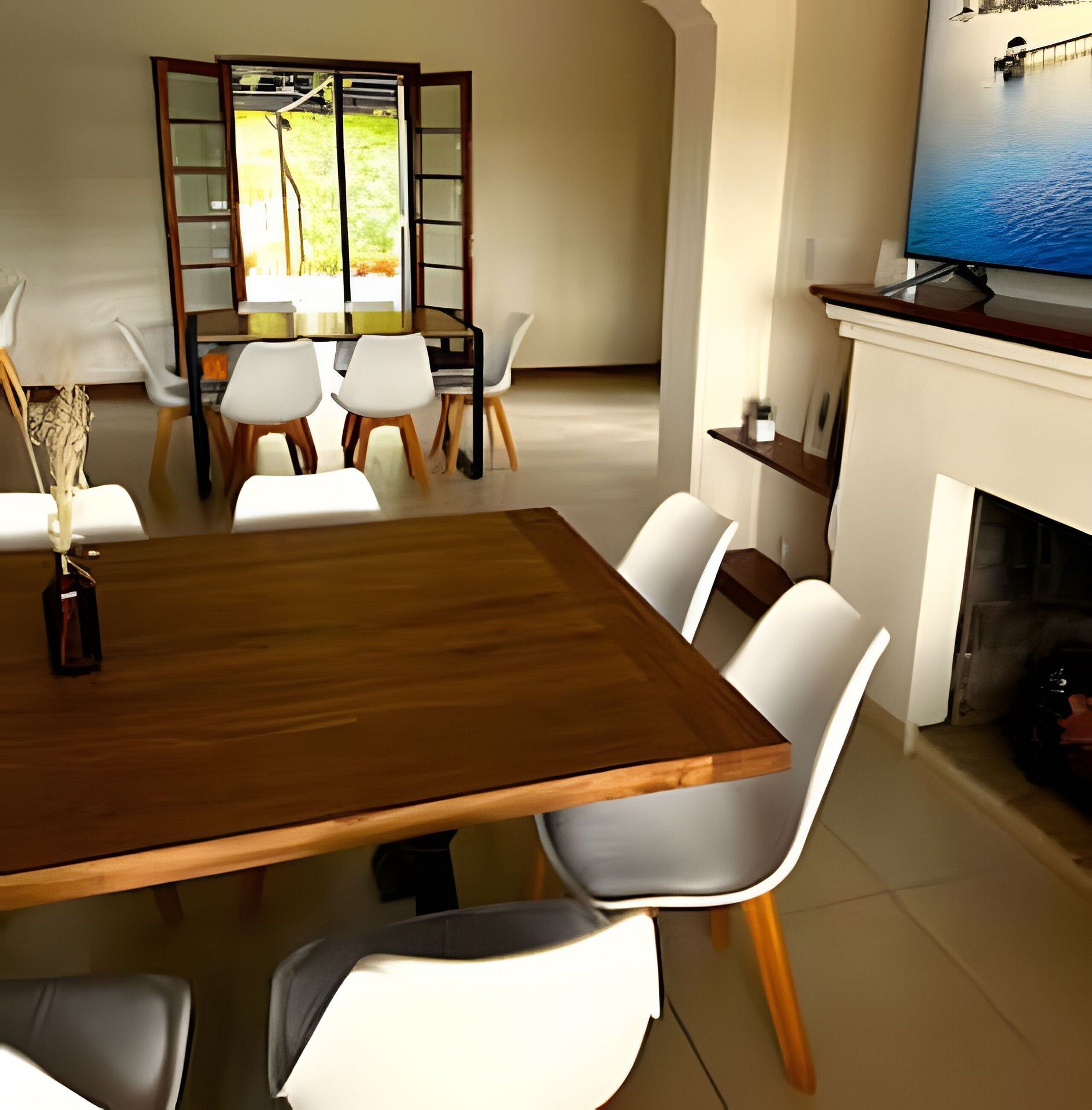 Un coin repas avec une table en bois, des chaises blanches, une porte vitrée donnant sur l’extérieur et une cheminée sur laquelle est accrochée une télévision.
