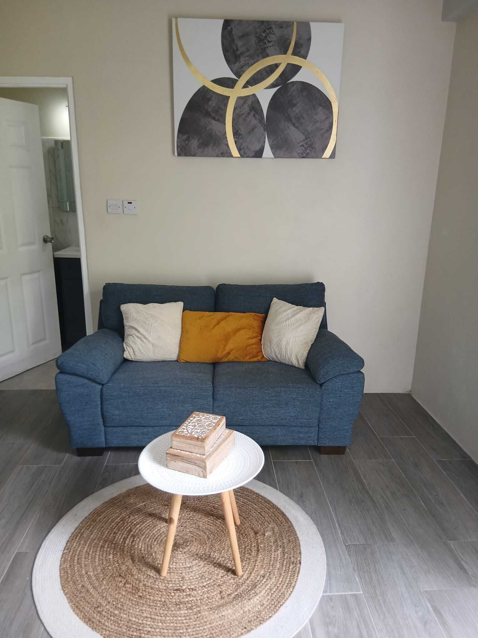 A living room with a blue sofa, a small white table with a woven rug, and a painting on the wall.