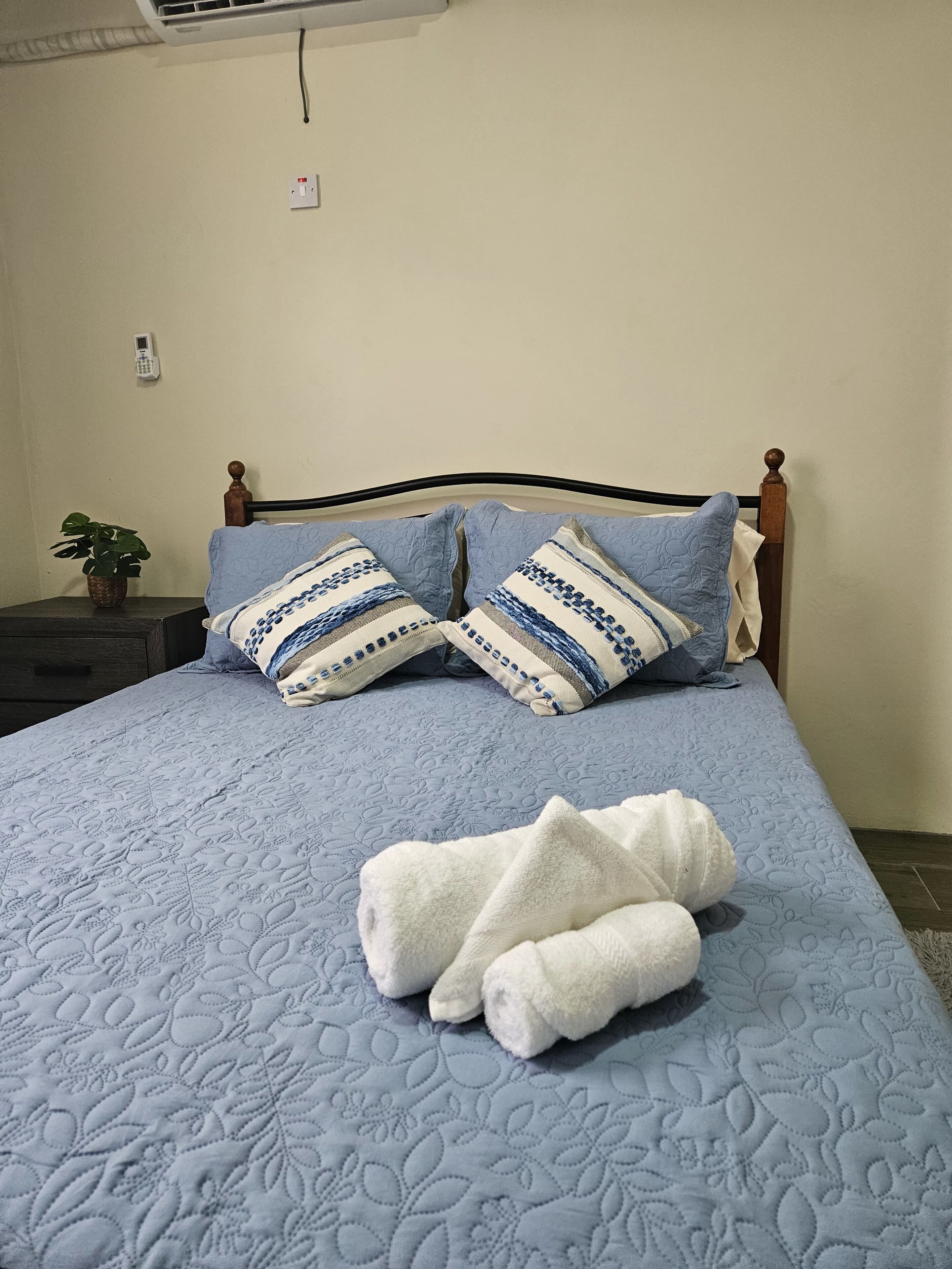 Una cama bien hecha con colcha azul, dos almohadas a rayas y dos toallas blancas dobladas.