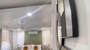 A bedroom with a bed, wooden flooring, a ceiling fan, and a wall-mounted air conditioning unit. - Cypress Suites (Roseau)
