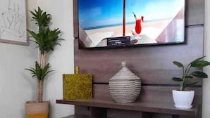 A modern living room with a flat-screen TV mounted on a dark wooden console, a potted plant, a decorative vase, and a patterned rug. - Cypress Suites (Roseau)