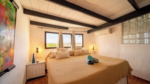 A bedroom with a large bed, two windows with curtains, a bedside table with a lamp, and a wall-mounted air conditioner. - Casa Natura 23 (San Bartolomé)