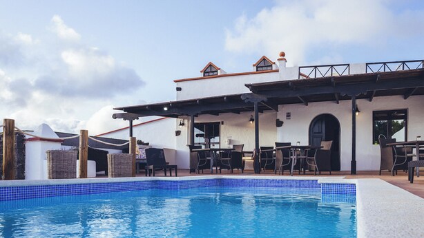 A pool area with a clear blue pool, outdoor seating, and a building with a white facade and red roof. - Casa Natura 23 (San Bartolomé)