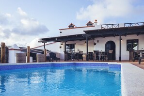 Un espace piscine avec une piscine à l’eau d’un bleu limpide, des sièges d’extérieur et un bâtiment à la façade blanche et au toit rouge.