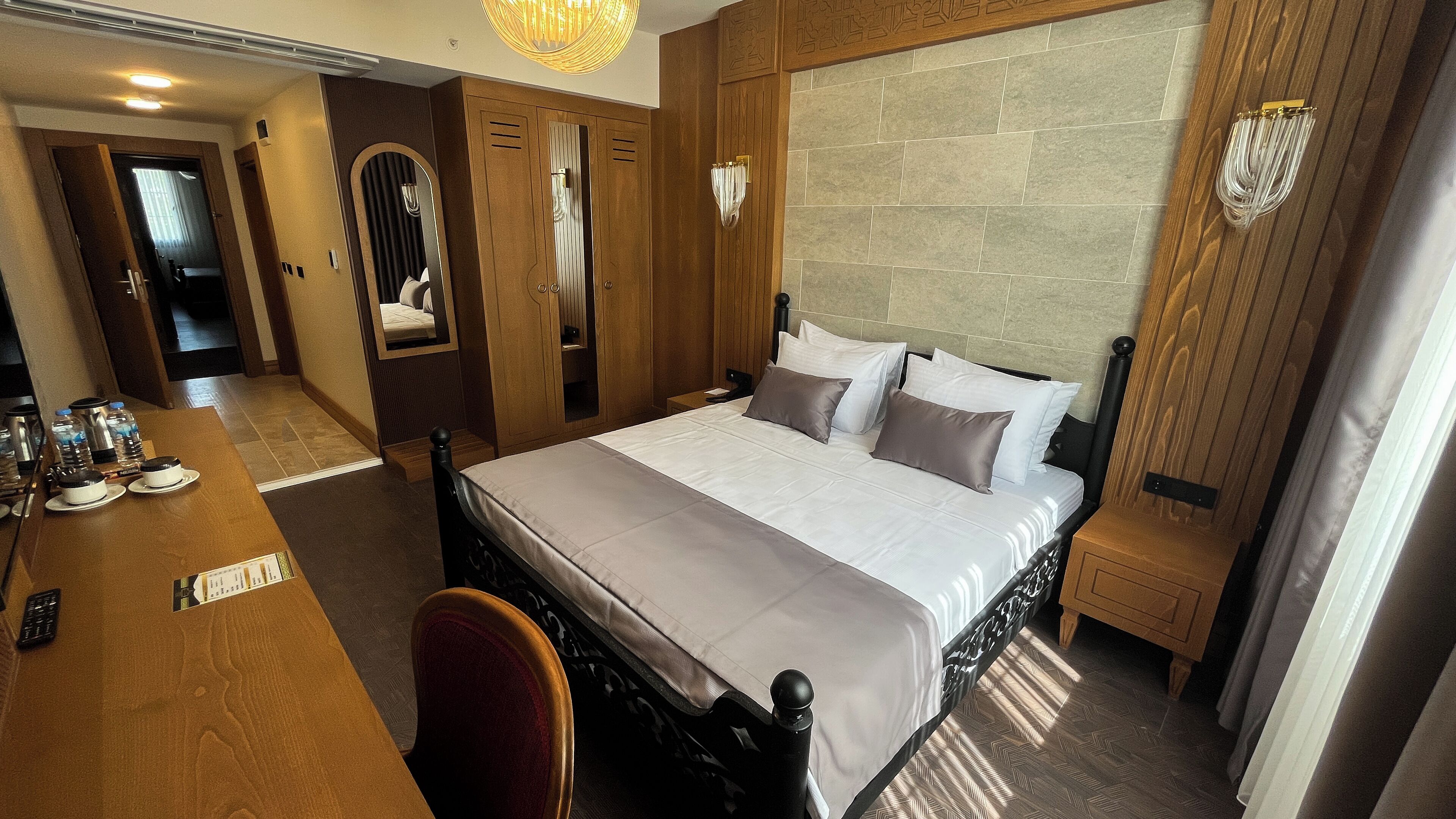 A hotel room with a large bed, wooden headboard, a bedside table with a lamp, a mirror, and a wooden desk with a chair and water bottles.