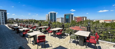 An outdoor terrace with tables and chairs, a tiled floor, and a view of city buildings in the background.