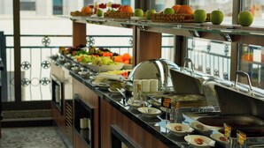 A buffet setup with various fruits and dishes, a tiled floor, and a view of buildings through large windows. - TURİSTİK PALAS OTEL (Diyarbakir)