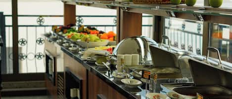A buffet setup with various fruits and dishes, a tiled floor, and a view of buildings through large windows.