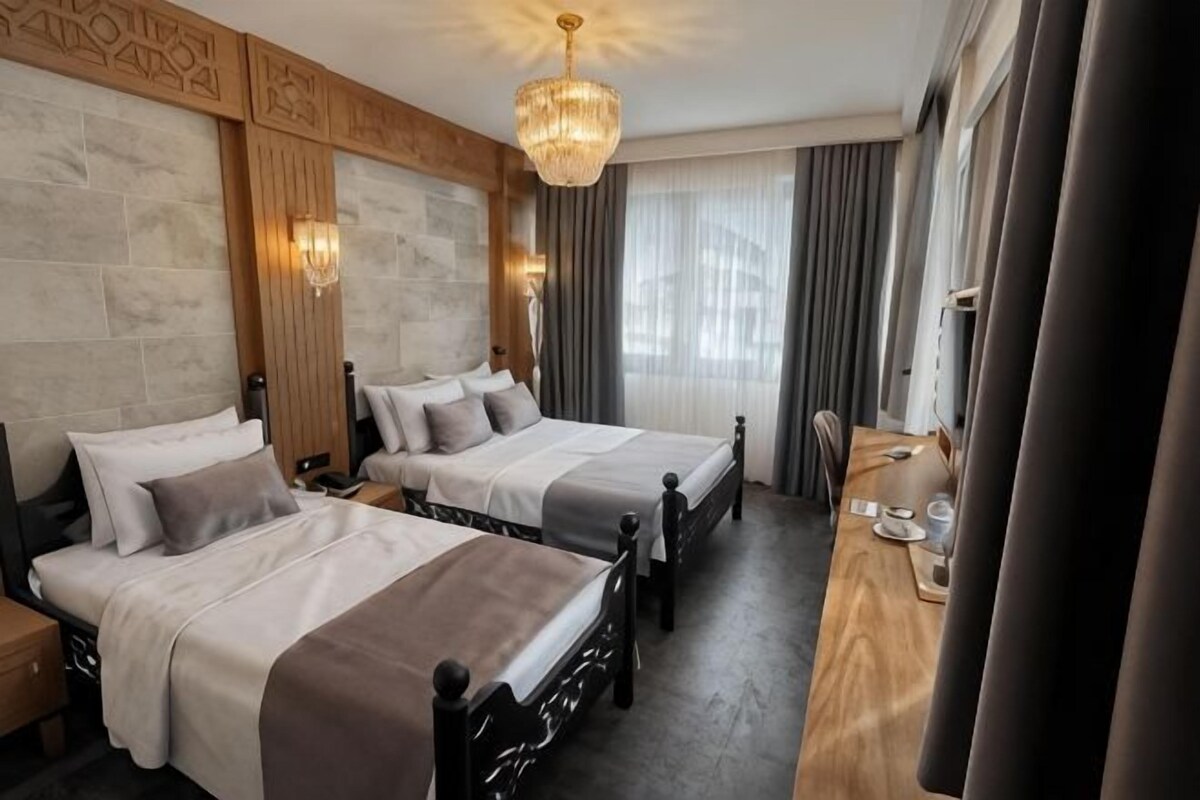 Camera d'albergo con due letti, una testiera in legno, un lampadario, una scrivania in legno e un'ampia finestra con tende.