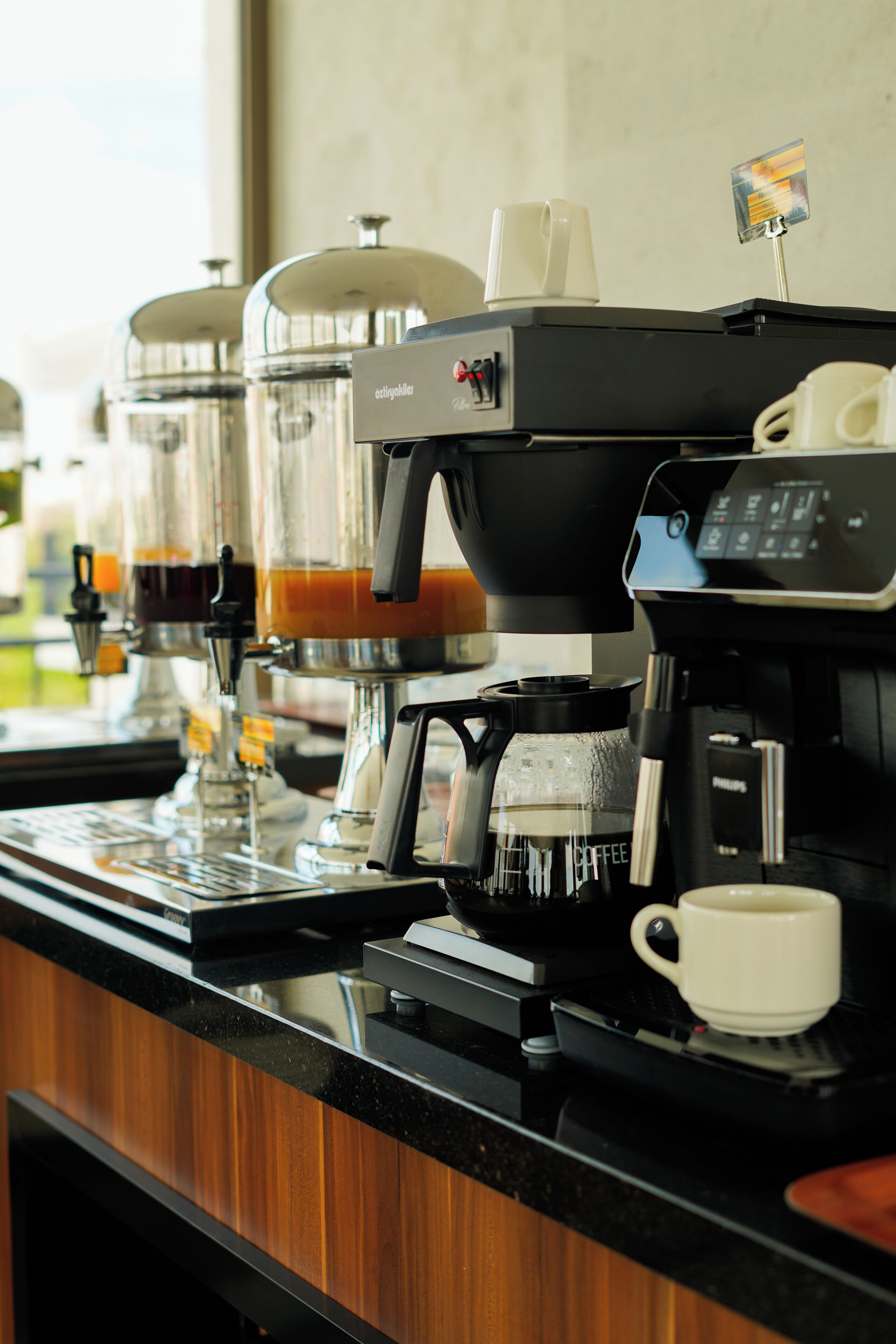 A coffee machine with multiple dispensers and a digital display, a white cup, and a menu card on the counter.
